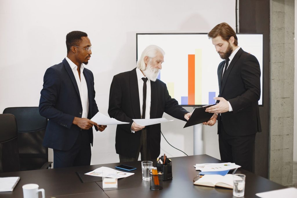 Three businessmen discussing documents in a conference setting with a presentation screen. — Enjoy the Latest Trends in Vapin