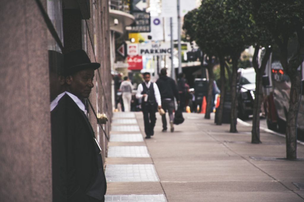 A man in a suit and hat leans against a city building, observing pedestrians. — Keys to Successfully Promoting Yourself as an