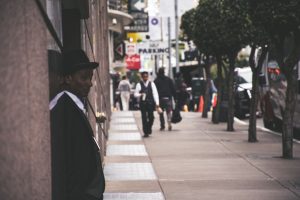 A man in a suit and hat leans against a city building, observing pedestrians. — Keys to Successfully Promoting Yourself as an