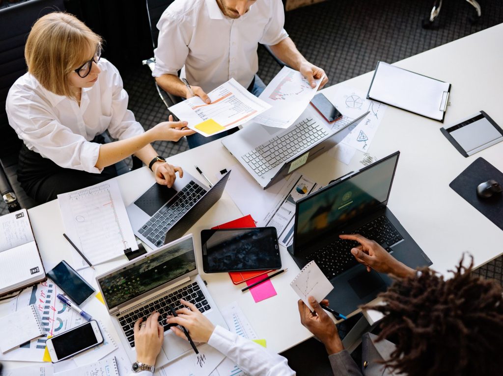 Top view of a diverse work team engaged in a collaborative office meeting, discussing papers and using laptops. — Search engi