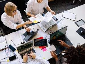 Top view of a diverse work team engaged in a collaborative office meeting, discussing papers and using laptops. — Search engi