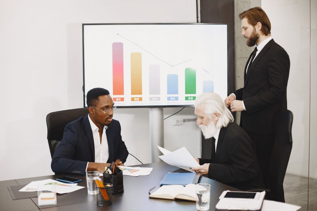 Three businessmen discussing financial data in a modern office setting with charts. — A Must For Your Financial Futur