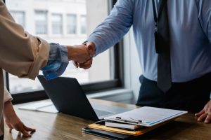 Crop unrecognizable coworkers in formal wear standing at table with laptop and documents while greeting each other before mee