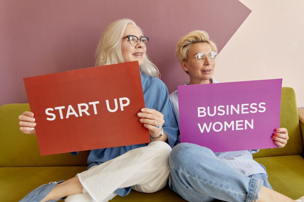 Two women on a sofa holding signs promoting startup and business women. — What are the Elements of a Resume Sample